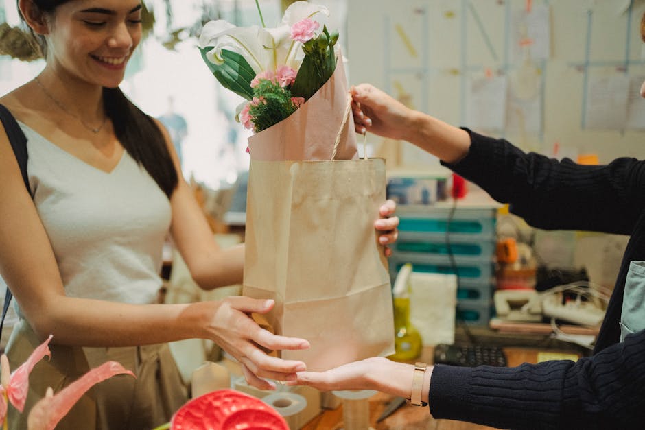 A woman receiving a floral gift from a flower shop assistant at flowerdeliverybaldock.co.uk. The bouquet features fresh white lilies and pink carnations arranged in pink wrapping paper. The assistant